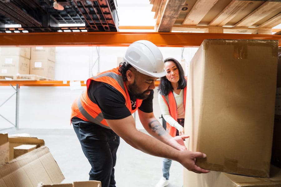 Two warehouse workers in orange safety vests, one wearing a white hard hat, work together to lift a large box from a shelf in a Warren MI storage area—always mindful of proper technique to help avoid shoulder pain on the job. Two warehouse workers in orange safety vests, one wearing a white hard hat, work together to lift a large box from a shelf in a Warren MI storage area—always mindful of proper technique to help avoid shoulder pain on the job.