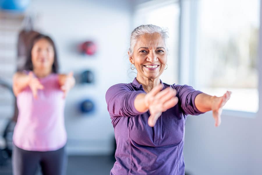 Two women are exercising indoors in Warren MI. The woman in the foreground smiles, wearing a purple long-sleeve shirt and extending her arms forward—a common stretch used in shoulder pain treatment. Another woman in pink reaches forward, slightly blurred. Two women are exercising indoors in Warren MI. The woman in the foreground smiles, wearing a purple long-sleeve shirt and extending her arms forward—a common stretch used in shoulder pain treatment. Another woman in pink reaches forward, slightly blurred.