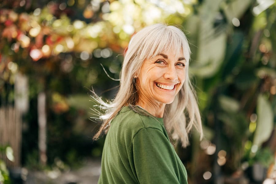 A smiling older woman with long gray hair and a green shirt stands outdoors in a sunlit garden, enjoying the vibrant foliage after receiving shoulder pain treatment in Warren, MI.