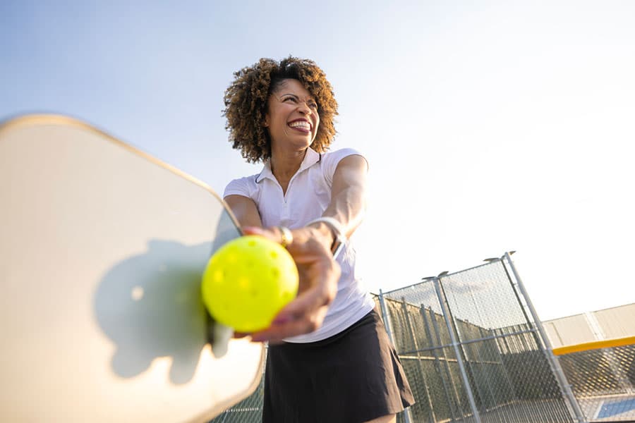 A woman smiles while playing pickleball outdoors in Warren, MI, holding a yellow ball and paddle near a chain-link fence on a sunny day—enjoying the game after finding effective shoulder pain treatment.