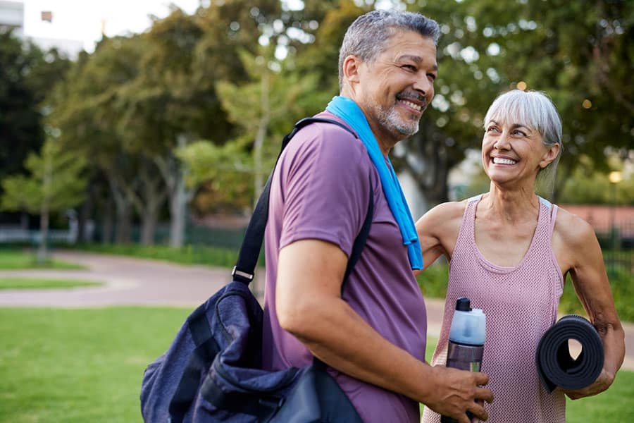 An older man and woman in athletic clothes smile at each other outdoors in a park near Warren MI. The man has a gym bag and towel; the woman holds a yoga mat and water bottle—perfect for their post-physical therapy routine. Trees and grass are in the background.