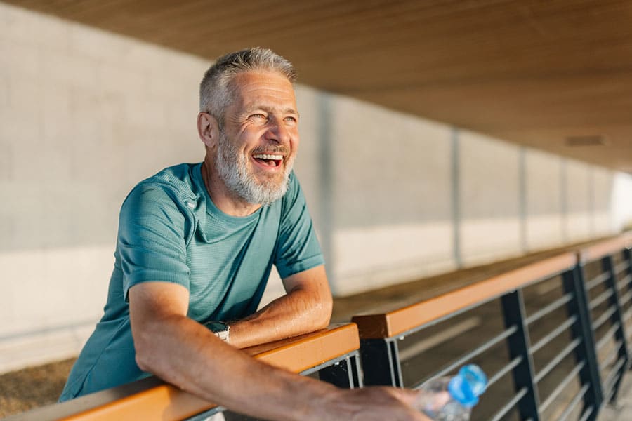 A smiling older man with gray hair and beard leans on a metal railing outdoors in Warren MI, wearing a teal athletic shirt and holding a water bottle, with sunlight illuminating his face—an inspiring image near a Physical Therapy Clinic.