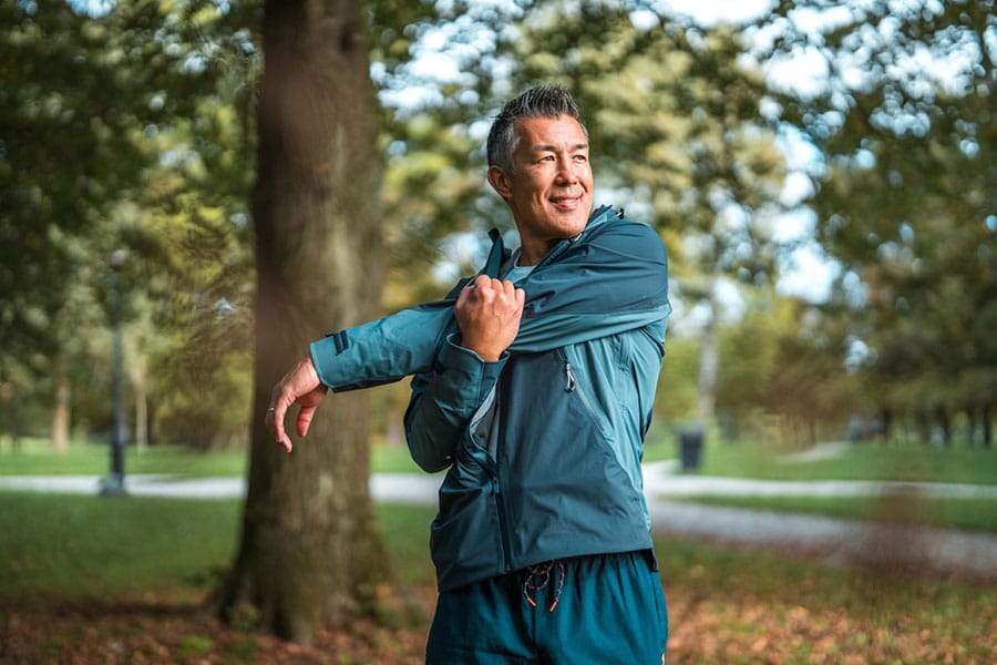 A man wearing a blue jacket stretches his arm across his chest while standing outdoors in a park, surrounded by trees and greenery, enjoying some gentle pain relief inspired by massage therapy techniques.