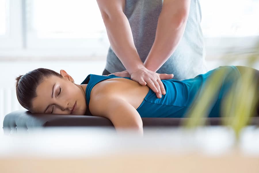 A woman in a blue tank top lies face down on a massage table with her eyes closed while a massage therapist applies pressure to her back for pain relief.