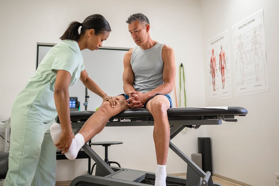 A physical therapist at a Warren MI Physical Therapy Clinic examines a man's leg as he sits on an exam table. The therapist gently holds and stretches his leg while he looks down, with anatomical charts visible on the wall.