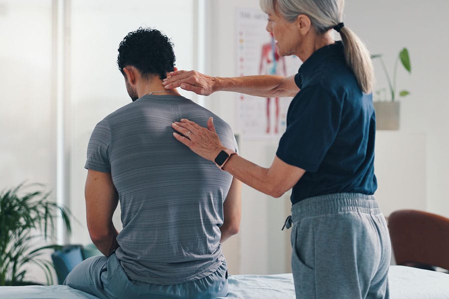 A physical therapist at a Physical Therapy Clinic in Warren MI examines the upper back and shoulder area of a seated man. The man faces away from the camera as the therapist gently touches his back in a clinic setting.