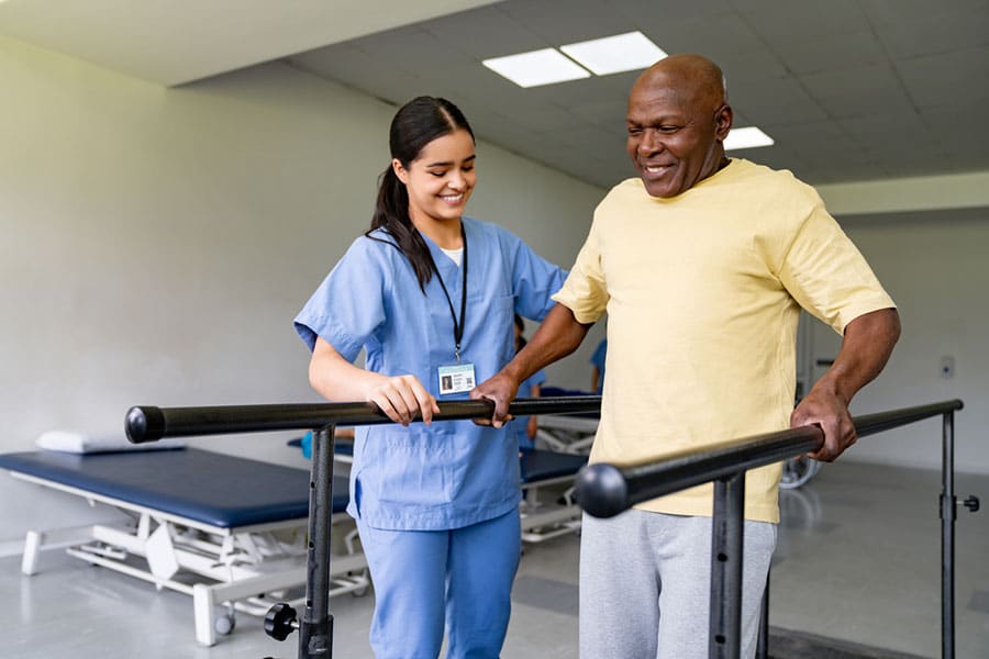 A smiling healthcare worker assists an older man as he walks between parallel bars in a Physical Therapy Clinic in Warren MI. The man holds the bars for support, with therapy beds visible in the background.