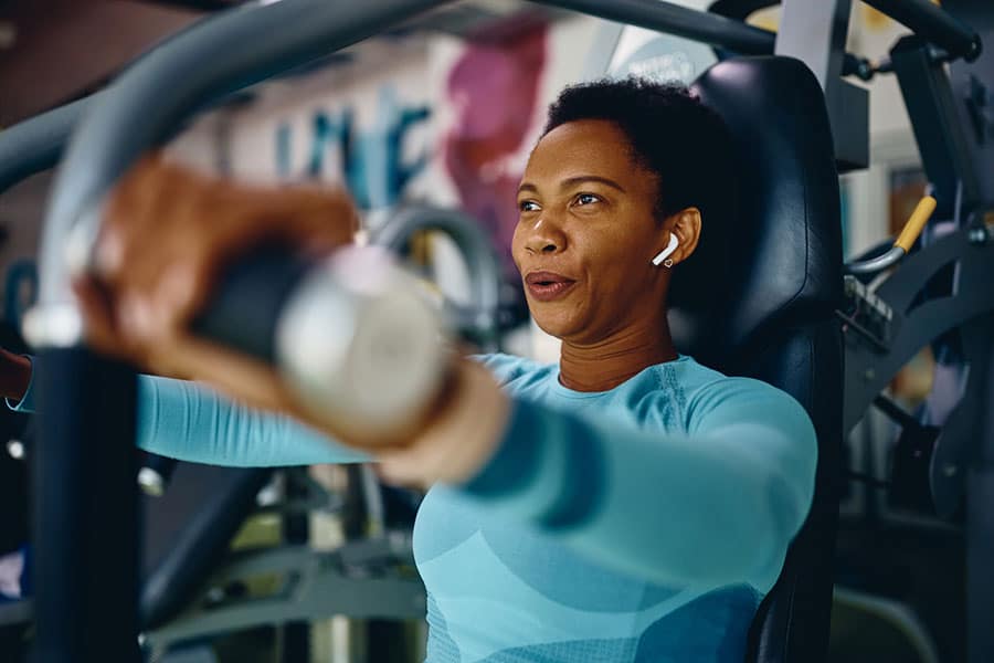A woman wearing wireless earbuds and a blue workout top uses a chest press machine at the gym in Warren MI, looking focused and determined—perfect inspiration for anyone visiting a local physical therapy clinic.