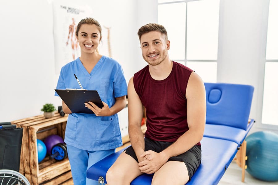 A woman in blue scrubs holding a clipboard stands next to a smiling man in athletic wear sitting on a medical exam table in a bright clinic room, highlighting their focus on recovery and pain relief.