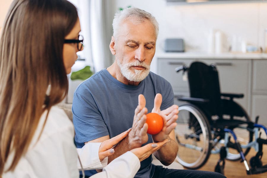 An elderly man holds a red ball with guidance from a female therapist, demonstrating the benefits of occupational therapy in regaining independence. A wheelchair is visible in the background in a bright, home-like setting.