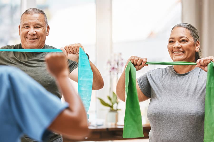 Two older adults smile while exercising with resistance bands indoors, following along with an instructor who faces away from the camera—a great example of independent seniors embracing steady aging through active living.