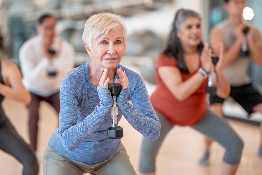 A group of adults, including a senior woman in front, exercise together indoors, holding dumbbells and performing squats—demonstrating the benefits of moving beyond joint degeneration and osteoarthritis through regular physical activity.