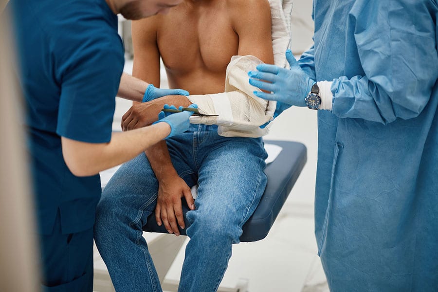 Two medical professionals in blue gloves and scrubs are assisting a seated, shirtless man by examining and supporting his bandaged arm in a clinical setting, providing attentive care as part of his post-injury recovery.