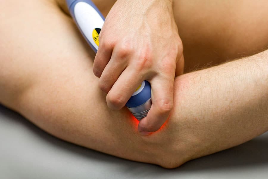 A person applies a handheld laser therapy device with a red light to their elbow, likely for pain relief, recovery, or the treatment of sports injuries.