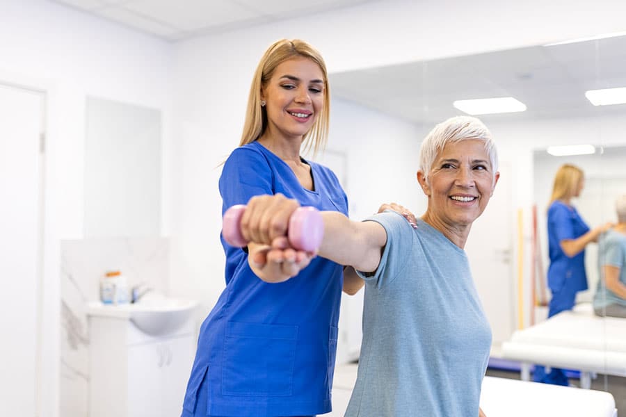 A healthcare professional assists a smiling older woman as she lifts a pink dumbbell in a bright clinic room, demonstrating how occupational therapy can make everyday tasks easier.