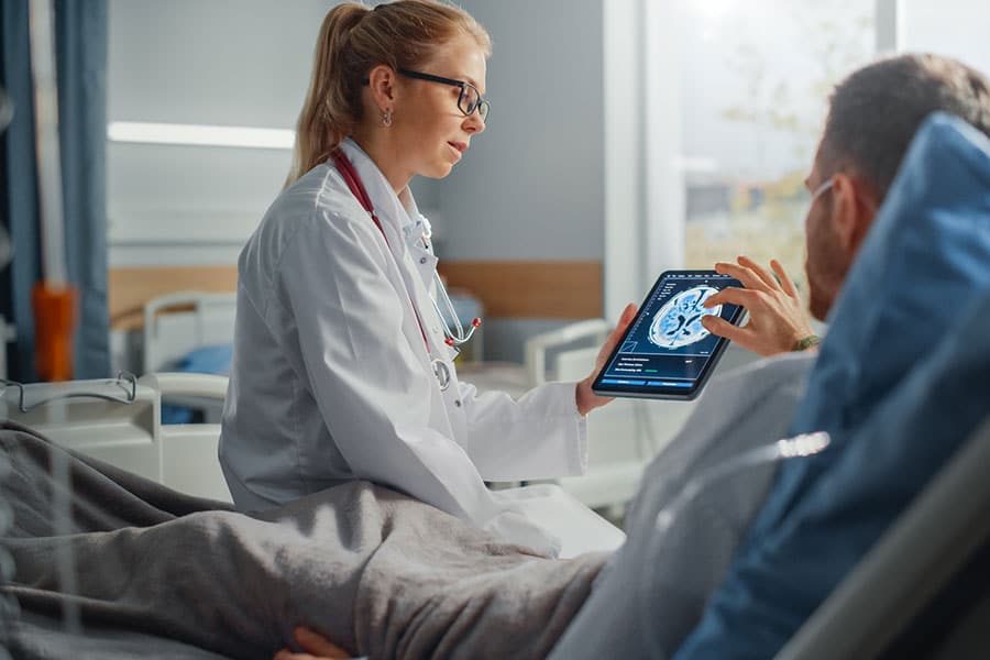 A doctor in a white coat shows a brain scan on a tablet to a patient in a hospital bed to discuss neurological recovery.