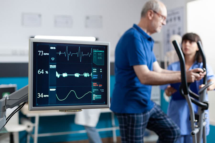 A man uses an elliptical machine during neurological rehabilitation as a clinician observes, with a monitor in the foreground showing his EKG and vital signs.
