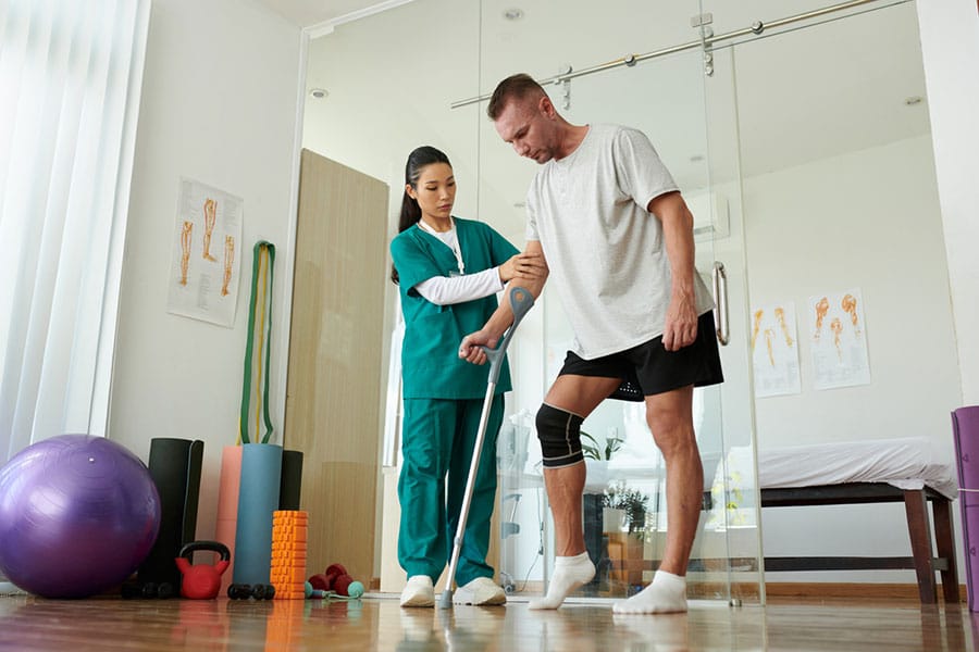 A physical therapist supports a man using a single crutch as he practices walking during neurological rehabilitation.